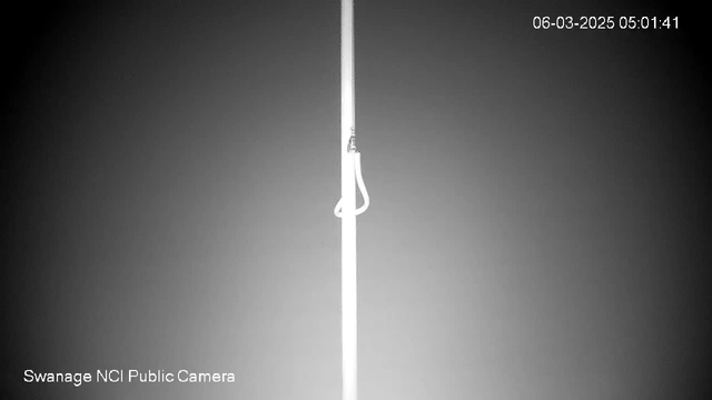 A tall, thin white pole stands vertically against a dark gradient background. A white looped rope is attached to the pole, near the top. The image is timestamped, showing the date and time in the upper right corner. The camera's label "Swanage NCI Public Camera" appears at the bottom left.