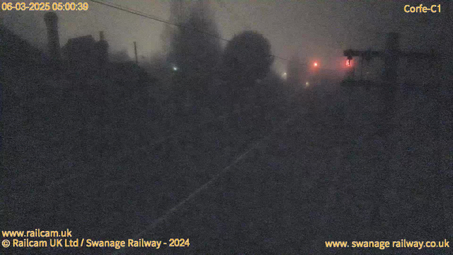 A dimly lit scene captured by a webcam, with heavy fog obscuring visibility. In the foreground, silhouettes of trees and structures are faintly visible. A few red lights can be seen in the background, possibly from signals or warning devices. The overall atmosphere is gloomy and dark, indicating that it is early morning, around 5:00 AM. The date and the camera's location are also noted at the top.