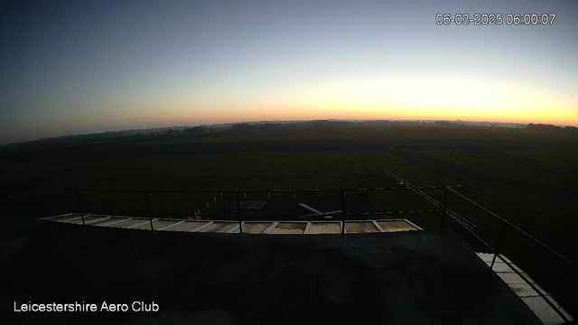 A wide view from a webcam at dawn, showing a clear sky transitioning from dark blue to soft orange and yellow near the horizon. In the foreground, a sloped roof with metal panels can be seen, and the ground below is flat, likely an airfield, with sparse vegetation. The scene conveys a serene early morning atmosphere, indicating the start of a new day. Text in the lower left corner reads "Leicestershire Aero Club," and the timestamp indicates the image was taken on June 3, 2025, at 6:00 AM.