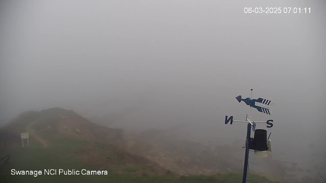 A foggy scene is depicted with poor visibility. In the foreground, a weather vane on a pole indicates direction, with the letters "N" and "S" visible. To the left, there is a sign partially obscured by fog, and the rugged coastline can be seen leading towards the sea. The background is filled with gray fog, obscuring any further details. The timestamp in the corner shows the date and time as 06-03-2025 07:01:11.