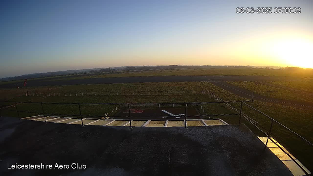 A sunrise scene viewed from a high vantage point of an airport. The sky transitions from deep blue to light orange near the horizon as the sun rises. In the foreground, a railing surrounds a flat rooftop, with the runway and grassy fields stretching out below. The runway is marked by white lines, and a small airplane is visible on the tarmac. There are several white fences bordering the grassy areas. The image captures the tranquility of an early morning at an aerodrome.