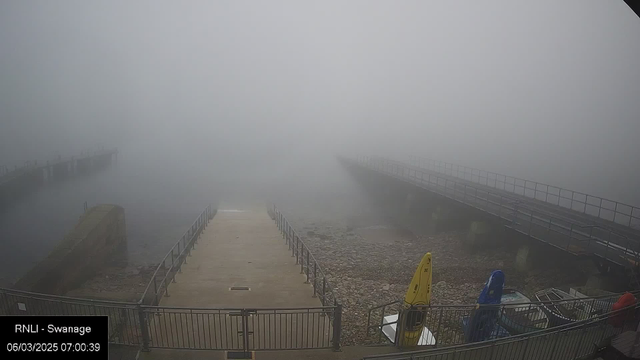 A foggy scene at a harbor with limited visibility. A wide, empty concrete path leads from the foreground to the water's edge. To the left, a rock structure is partially visible. In the bottom right corner, there are several brightly colored kayaks, including a yellow one, resting on a metal platform. A long pier extends to the right, disappearing into the fog, while a railing lines the pathway and pier. The overall atmosphere is misty and obscured, creating a sense of calm and quiet.