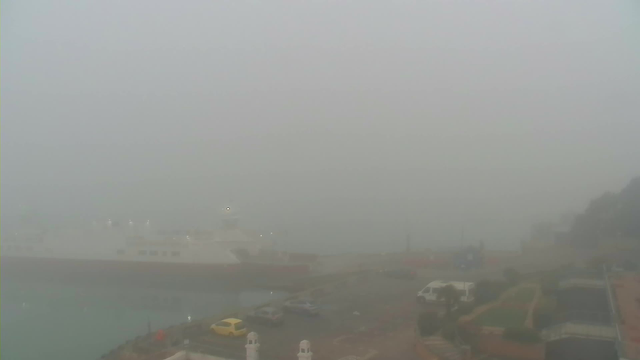 A foggy view of a harbor area, with very limited visibility. A large white ferry is partially visible on the water, and several vehicles are parked on the road, including a yellow car. The surrounding area is obscured by thick fog, making details indistinct, with hints of buildings and greenery in the foreground.