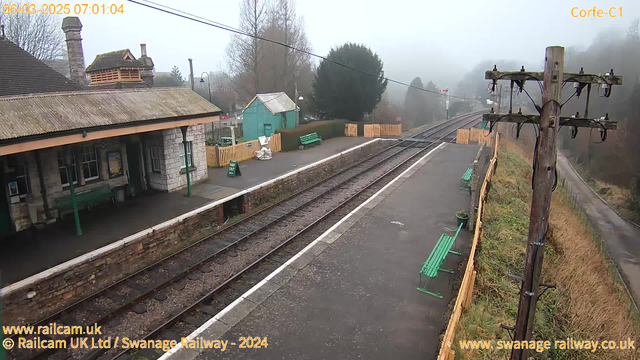 A foggy scene at a railway station. On the left, a brick building with a slanted roof and green benches outside. A tall wooden utility pole with wires stands in the foreground. The railway tracks run parallel to the platform, which is lined with a wooden fence on the right. A small green shed can be seen in the background, and the surrounding area appears lush with fog obscuring distant views. The overall atmosphere is calm and quiet, typical of an early morning.