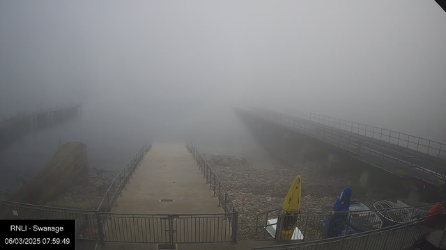 A foggy scene at a dock. The view shows an empty jetty extending into the mist, with a concrete pathway leading to it. On the left, a rocky shoreline is visible, and on the right, several brightly colored kayaks are stored. The visibility is very low due to the thick fog, creating a quiet and subdued atmosphere.