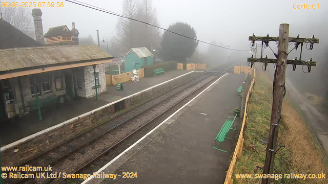 A foggy railway station scene shows a platform with a few green benches and a stone building with a sloped roof on the left side. The area is surrounded by trees, and a wooden fence is positioned in the background. Electric poles with wires line the area, leading towards the tracks that are visible on the right. Visibility is low due to fog, creating a muted atmosphere. The date and time are displayed at the top of the image.