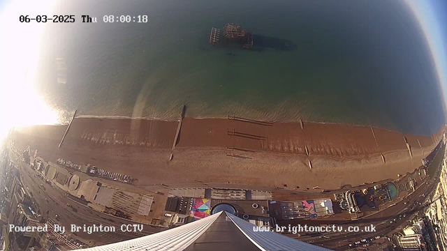 A panoramic view from a high perspective looking down at a beach along the coastline. The image captures the sandy beach with several wooden piers extending into the water. The sea is in shades of blue and green, reflecting sunlight. There are structures along the beach, including umbrellas and a few vehicles visible on a nearby road. The date and time are displayed in the top corner, indicating it is early morning on March 6, 2025.