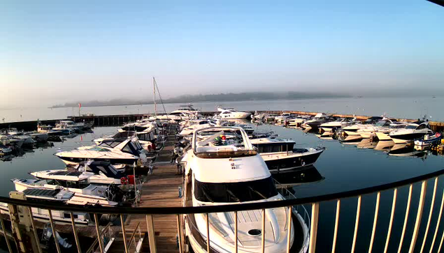 A calm marina scene at dawn, showing numerous boats docked at a wooden pier. The water is still and reflects the boats and the clear blue sky above. Light fog can be seen in the distance, adding a serene atmosphere to the setting. The boats are a mix of sizes and designs, mostly sleek with white and dark colors, some featuring vibrant accents. A small red flag is visible in the background near the marina entrance.