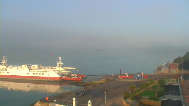 A ferry is docked at a harbor during a calm morning, with a foggy atmosphere. The water is still, reflecting the ferry and nearby structures. The foreground shows a parking area, marked with a few vehicles and traffic signs. Surrounding the parking lot are trees and landscaped greenery. In the background, there are buildings and a hazy shoreline. A red buoy is visible on the water.