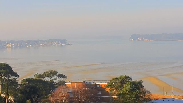 A serene waterfront scene featuring calm blue waters reflecting a pale sky. In the background, a line of boats is docked, and a faint outline of land can be seen, likely with trees and buildings. The foreground shows a mix of green trees and a few buildings, with some structures visible just above the tree line. Light fog or mist adds a soft atmosphere to the scene, suggesting a tranquil, early morning setting.