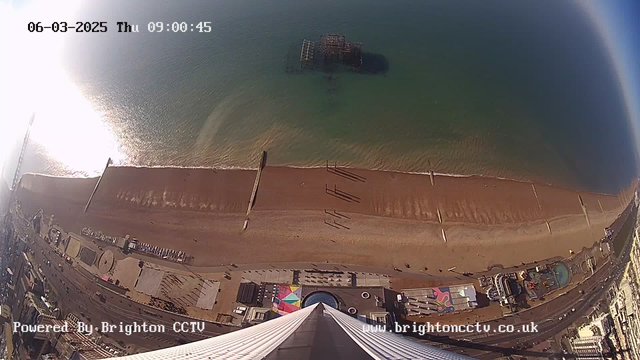 Aerial view of a sandy beach along a coastline, with clear blue water gently lapping at the shore. The lower half of the image shows a wide beach area with wooden posts and shadows cast by them. To the left, part of a pier extends into the water. In the foreground, there are colorful structures, possibly beach huts or amusement rides. The image captures a sunny day with bright sunlight reflecting off the water surface. The date and time displayed on the top of the image indicate it is March 6, 2025, at 09:00:45.