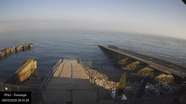 A calm seaside view shows a wooden walkway extending into the water from a stone platform. The water is smooth and reflects the sky, with a light mist in the distance. To the right, there are two yellow kayaks and a blue boat next to the shore, surrounded by small rocks and pebbles. The scene has a serene atmosphere, with no people visible. The time displayed is 09:00 on March 6, 2025.