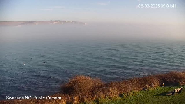 A coastal scene showing calm waters with a slight mist hovering over the surface. In the background, low cliffs are partially obscured by fog. There are some small white buoys floating on the water. In the foreground, there is a patch of green grass and a couple of benches. The atmosphere is serene, with blue sky visible above.