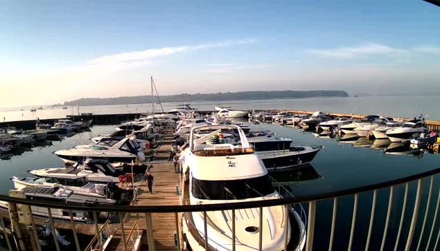 A marina filled with numerous boats of various sizes, docked along wooden piers. The water is calm and reflects the clear blue sky above, with a few wispy clouds. In the distance, a shoreline is visible, and a person is walking along the dock. The scene conveys a tranquil morning atmosphere.