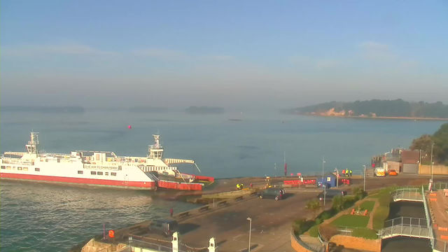 A ferry is docked at a harbor on calm waters, with a clear blue sky above. The ferry is white with a red bottom and has multiple decks. Near the ferry, several people in bright clothing are gathered on the dock. In the background, there are small boats and islands visible across the water, while a coastal area shows trees and sandy shores. The scene is bright and serene, suggesting a peaceful morning.