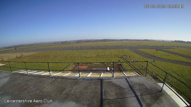 A clear sky with bright sunlight above a grassy field and paved runways. In the foreground, there is a railing and a small helipad marked with a white rectangular shape. The field stretches out in the distance, with a fence outlining the area. There are no visible aircraft. The image captures a tranquil, open space typical of an aerodrome.