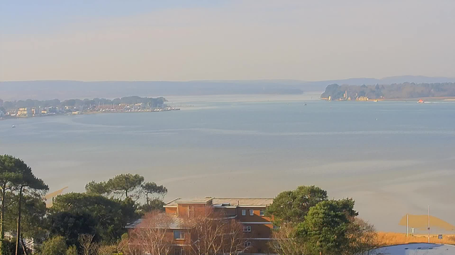 A view of a tranquil body of water, with soft waves and a calm surface reflecting light. In the foreground, there are several trees and a building with a flat roof. In the background, a shoreline is lined with small boats and a cluster of houses. Beyond the shore, there are rolling hills fading into the distance, partially obscured by a gentle haze. The sky is bright and clear, suggesting a pleasant day.