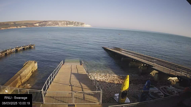 A view of a calm seaside scene with clear blue water and a sandy beach. In the foreground, there are two piers extending into the water. One pier is straight and wide with a railing, leading down to the water. The other pier is narrower and slightly angled. On the right side, there are several boats, including yellow and blue kayaks, resting on a rocky shore. In the background, cliffs are visible along the shoreline, with greenery at the top. The sky above is clear and bright. A timestamp is displayed in the lower left corner.