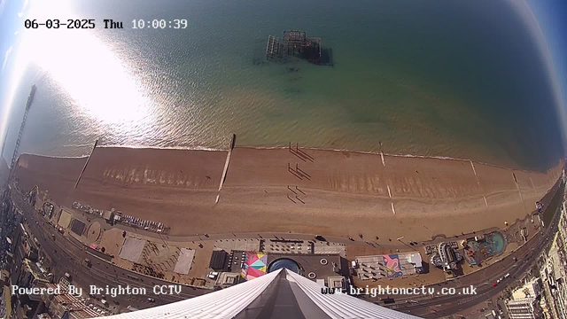 An aerial view of a beach scene during the day, with the shoreline stretching across the bottom of the image. The sand is a light brown color, showing some patterns and footprints. The water is a gradient of blue and green, reflecting sunlight. There are several people walking along the beach, looking small against the expansive shoreline. A pier extends into the water on the left side, and an old, partially submerged structure is visible in the water. Below, various colorful attractions and structures are set up along the beachfront, indicating a lively area. The top of the image has a clear blue sky with a few scattered clouds. Time and date information is displayed in the upper left corner.