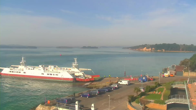 A white and red ferry is docked at a quiet harbor, surrounded by calm blue water. In the background, small islands are visible under a clear sky. To the right, a sloping beach with a light-colored cliff and greenery can be seen. Several parked vehicles are lined up on a paved area near the ferry terminal, with a few people walking nearby.