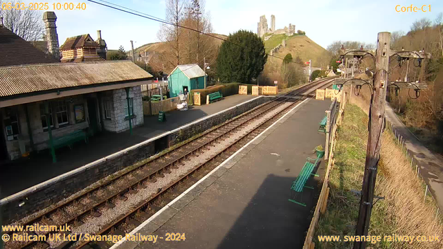 A narrow-gauge railway station platform is depicted, consisting of stone and concrete. Several benches are visible along the platform, with the foreground showing railway tracks leading off into the distance. In the background, there is a small green shed and a wooden fence, along with a backdrop of rolling hills and a castle ruin on the hilltop. The sky is blue with light clouds, indicating a clear day. A telephone pole with wires stands to the right side of the image.