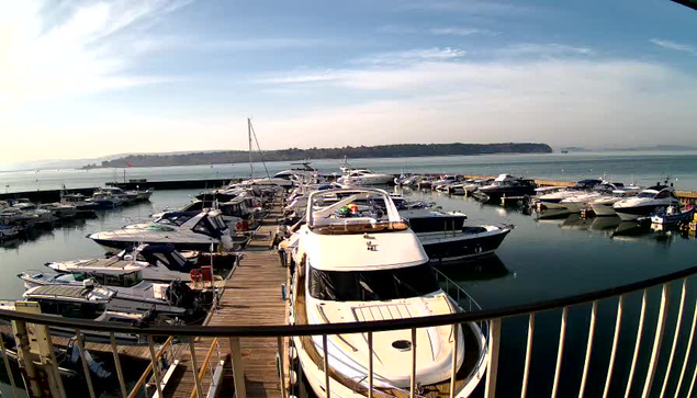 A view of a marina filled with numerous boats docked along wooden piers. The water is calm, reflecting the sky, and there are distant hills in the background. The sky is partly cloudy with a gentle brightness indicating daytime. The boats vary in size and shape, with some featuring canopies and visible details like navigation equipment. A few flags are seen in the distance, fluttering lightly in the breeze.