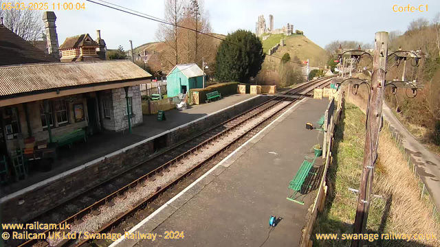 A railway station scene featuring a stone building with a sloped roof on the left, surrounded by trees. To the right, the tracks run straight with no trains in sight. A green shed is visible in the background, along with several green benches positioned along the platform. A wooden fence separates the platform area from a path lined with grass. In the distance, a hill can be seen topped with ruins of a castle, under a clear blue sky. The image is well-lit, suggesting a sunny day.