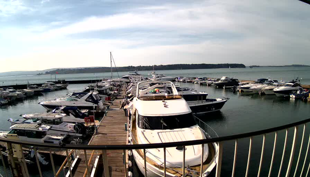 A view of a marina filled with various boats and yachts docked along wooden piers. The scene is bright with a clear sky and gentle waves. In the background, a green landscape is visible, alongside more boats on the water. The image captures a calm and sunny day.