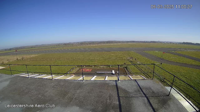 A clear blue sky is visible above a wide, open grassy field. In the foreground, there is a railing and a flat surface, suggesting a rooftop or balcony area. The ground is mostly green grass, with a few paved areas and a marked runway. A small red marker can be seen in the distance, possibly indicating a location on the runway. The overall scene is bright and sunny, showcasing a peaceful and spacious outdoor environment.