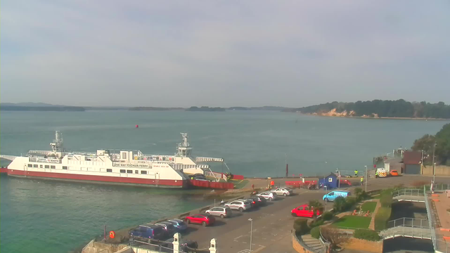 A ferry is docked at a terminal on a calm body of water, surrounded by distant islands and a cloudy sky. In the foreground, several parked cars are visible, including red and white vehicles. A few people are near the ferry and the terminal buildings, which have greenery and walkways. The landscape includes a shoreline with trees and rocky areas in the background.