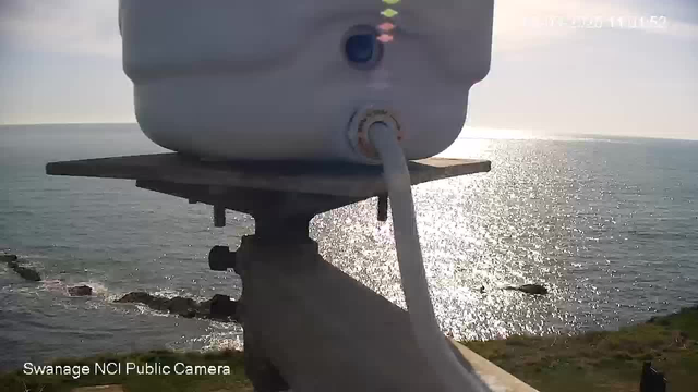 A white cylindrical container is positioned on a metal support. The container has a visible blue opening and a connected white tube. In the background, the sea sparkles under sunlight, with gentle waves and some rocks visible along the shoreline. The sky is clear with a hint of clouds. The image is taken from a public webcam.