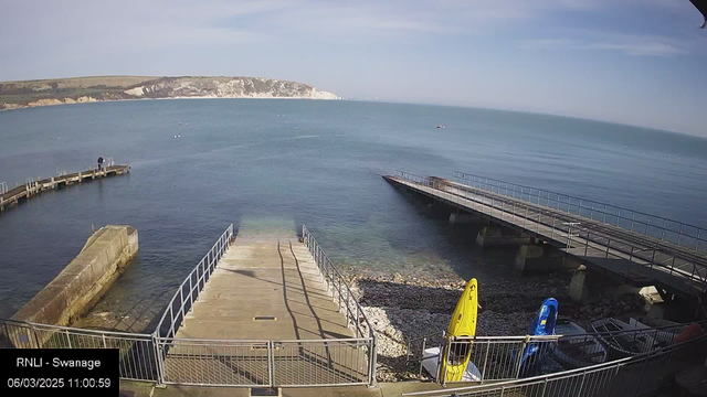 A view of a calm sea with a distant coastline featuring cliffs. In the foreground, there are two kayaks, one yellow and one blue, positioned on a small rocky beach next to a wooden ramp leading to the water. A wooden pier extends into the sea, partly visible on the left side of the image. The sky is clear with some soft clouds, creating a bright atmosphere. The time and date are displayed at the bottom of the image.