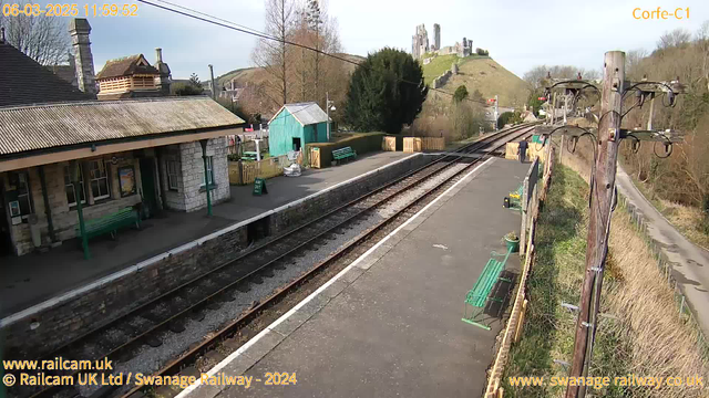 A view of a railway station with a stone building featuring a sloped roof on the left side. There are several green benches along the platform, and a light blue shed nearby. The tracks extend into the distance on the right, with a hill in the background topped by ruins and greenery, partially obscured by trees. The sky is clear with some clouds, indicating daytime. A telephone pole is present on the right, with wires extending across the top of the image.