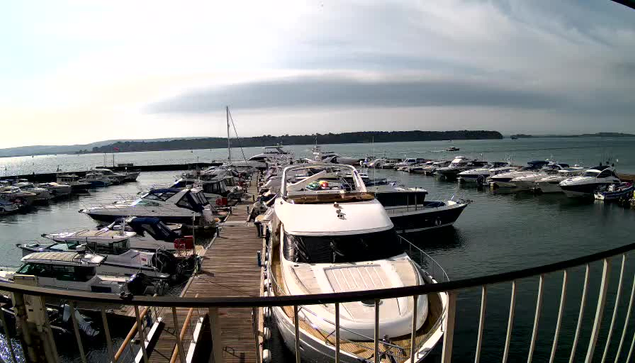 A view of a marina filled with numerous boats docked along a wooden pier. The foreground features a large yacht on the left, with its deck visible. Several smaller boats are tied up along the pier, and the water is calm, reflecting the light. In the background, there are more boats scattered across the water, with a hazy horizon and a mix of clouds above. The setting appears tranquil and sunny.