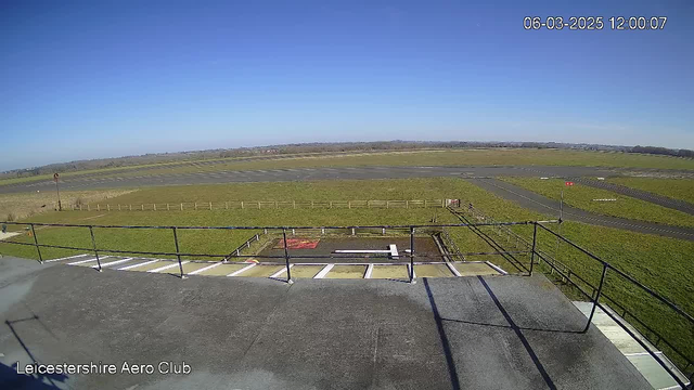 A wide view from a rooftop showing a clear blue sky above a grassy airfield. There are runways marked with white lines on the ground. In the foreground, a railing is visible along the edge of the rooftop. The grass is lush and green, and a few trees can be seen in the distance. No aircraft are visible on the runway. The time and date displayed at the top indicate it is noon on March 6, 2025.