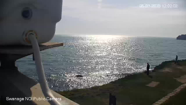 A view of the ocean with sunlight reflecting off the water, creating a shimmering effect. In the foreground, there is a small white structure, possibly part of the camera setup. To the right, two figures stand on the grass near the shoreline, with gentle waves lapping at the coast. The sky is mostly clear with some light clouds. The image captures a peaceful seaside scene.