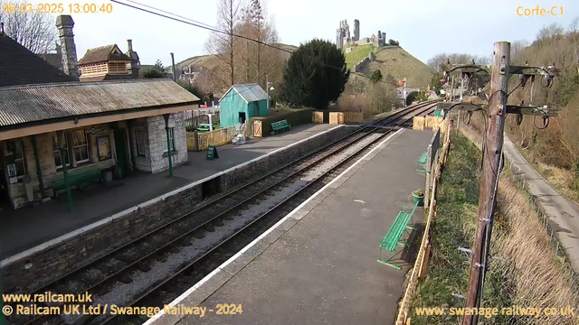 A view of a train station platform with green benches lining the side. The station's building, made of stone with a slate roof, is on the left, featuring large windows and a sign indicating "Cafe." In the background, there is a small wooden shed painted green, surrounded by a wooden fence. The railway tracks are visible, leading towards mountains with a castle ruin on top in the distance. The scene is bright with trees appearing bare, indicating early spring. There are utility poles with wires on the right side of the image, and a pathway leading into the countryside is visible towards the far right.