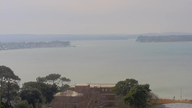 A wide view of a calm body of water with soft ripples, surrounded by trees on the left and a distant shoreline on the right. The sky is pale and hazy, casting a muted light over the scene. In the foreground, there is a partially obscured building with a flat roof, and some foliage surrounding it. Further out, the shoreline shows faint outlines of boats and structures along the water's edge.