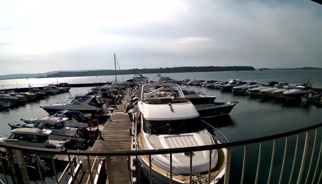 A view of a marina with numerous boats docked at different angles. The foreground features a large white yacht with a sleek design, while several smaller vessels surround it. The water is calm and reflects the overcast sky. In the background, land is visible along the horizon, and the scene is framed by a wooden walkway leading towards the marina.