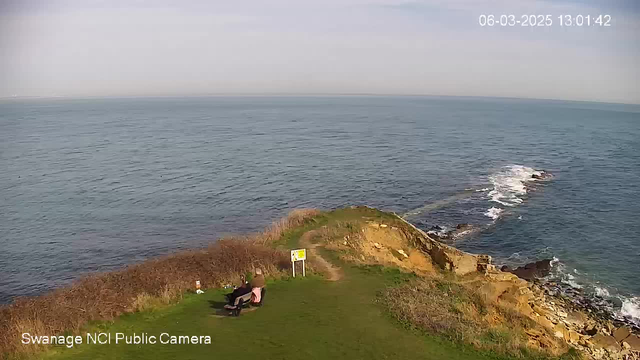 A scenic view of the coastline with calm blue waters extending to the horizon. In the foreground, a person sits on a bench on a grassy area overlooking the sea, near some low bushes. A sign is visible nearby, indicating a warning or information. The land gently slopes towards rocky outcrops where waves break against the rocks. The sky is mostly clear with light clouds.