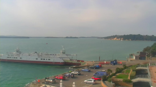 A ferry docked at a harbor, with its bow facing the shore. The water is calm and turquoise, with distant land visible in the background, featuring trees and a sandy shoreline. Various parked cars are lined up in a parking area to the right, and there are some people visible near the ferry. The sky is mostly clear with a hint of clouds.