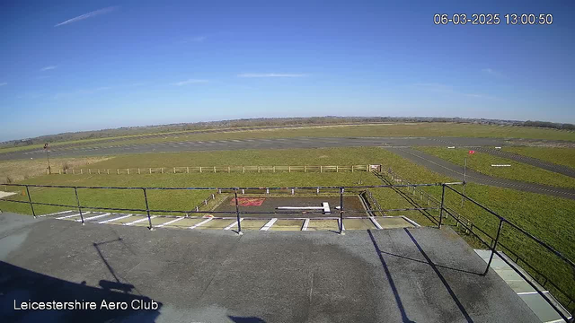 A clear blue sky is visible, with a few wispy clouds. Below, there is an expansive green field with a tarmac airstrip. The runway is marked with white lines and has a red windsock on one side, indicating wind direction. There is a railing in the foreground, likely from a viewing platform or balcony. The scene depicts a tranquil atmosphere at Leicestershire Aero Club on a sunny day.