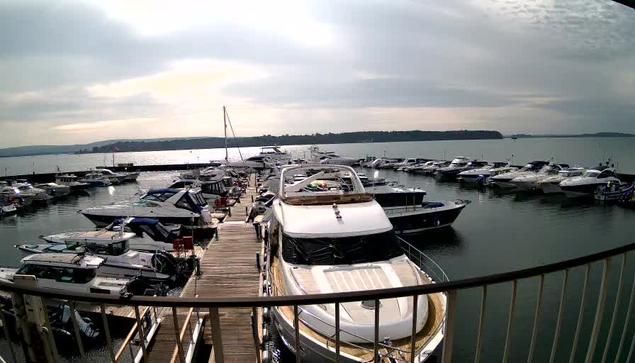 A view of a marina filled with boats, including several yachts and smaller vessels, docked along wooden piers. The water reflects the cloudy sky, creating a tranquil atmosphere. In the background, a distant shoreline is visible, with gentle hills rising behind the harbor. The scene conveys a calm, overcast day by the water.