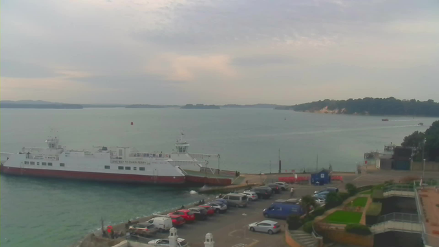 A view of a calm sea under a cloudy sky. On the left, a large white and red ferry is docked at a pier, with the word "FERRY" visible on its side. The water is dotted with a few small boats and a red navigational buoy. On the right, a parking area is filled with various cars, and behind it, there are some green walkways and landscaped areas. In the background, a distant island is visible with trees and a rocky shoreline.