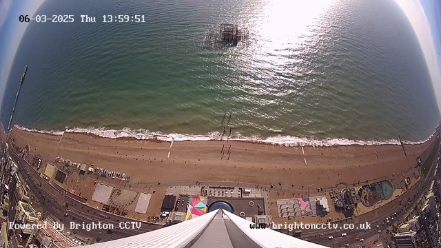 Aerial view of a beach in Brighton, showing golden sand lined with gentle waves, transitioning into a calm turquoise sea. In the distance, a partially submerged pier is visible. The shoreline features a variety of structures and colored canopies, while people can be seen walking on the beach. The scene captures sunlight shimmering on the water's surface, creating a bright and vibrant atmosphere.