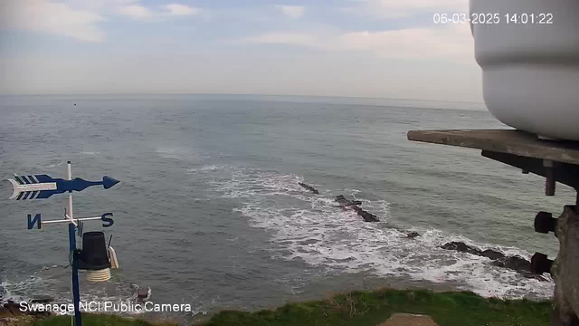 A view of the sea from a high vantage point. In the foreground, there is a blue and white weather vane pointing north, with a circular white object on the right side, possibly a container. Below the weather vane, there is a dark structure on the shoreline. The water is calm with gentle waves and some rocky formations are visible emerging from the sea. In the background, the sky appears partly cloudy with soft clouds.