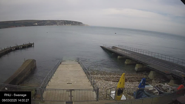 A view of a calm sea along a shoreline, with a concrete ramp leading down to the water on the left. A small, rocky beach is visible alongside the ramp. To the right, there are two piers extending into the water, both featuring railings. There are several boats stored nearby, including a yellow kayak, and the landscape in the background includes a green hillside and white cliffs under a cloudy sky. The image is timestamped March 6, 2025, at 14:00:27.