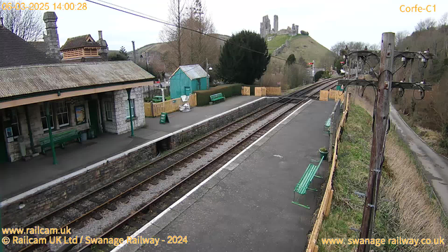 A railway station with a stone building featuring a sloped roof is visible in the foreground. There are green benches along the platform and a wooden fence surrounding the area. To the left, a small turquoise shed can be seen, and a sign reading "WAY OUT" is placed on the ground. In the background, a hill rises, topped with ruins, which are the remnants of a castle. The railway tracks run parallel to the platform, leading away from the station. The scene is set in a natural landscape with trees and grass lining the area.