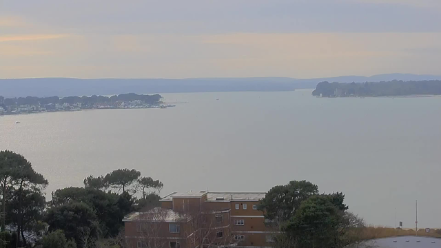 A serene view of a wide body of water, reflecting soft light under an overcast sky. In the distance, low hills are visible along the horizon, while the shoreline on the left features small boats and buildings. Closer to the foreground, there are clusters of trees and a brown building with multiple windows, surrounded by greenery. The overall atmosphere is calm and tranquil.