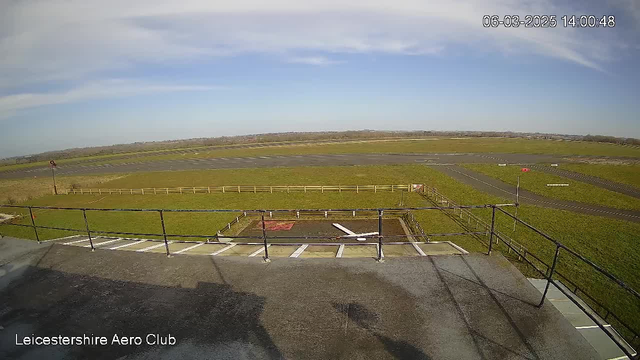 A view from a high vantage point overlooking an aerodrome. In the foreground, there's a flat rooftop area with a railing. Below, a patch of grass bordered by a wooden fence can be seen, leading to a long, paved runway that stretches across the middle of the scene. A small red indicator and white markings are visible on the runway. The sky is mostly clear with a few clouds, and the surrounding landscape is flat with sparse vegetation.